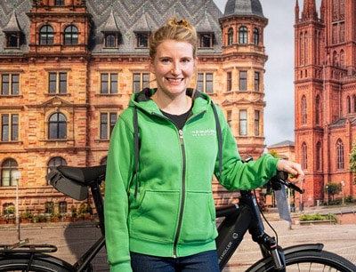 Julia aus Wiesbaden vor einer Wand mit Gebäude als Hintergrund lehnt an e-Bike