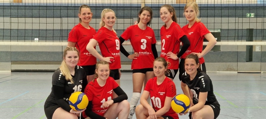 Damen Volleyballmannschaft mit roten Trikots und Volleybällen, stehen vor einem Volleyballnetz