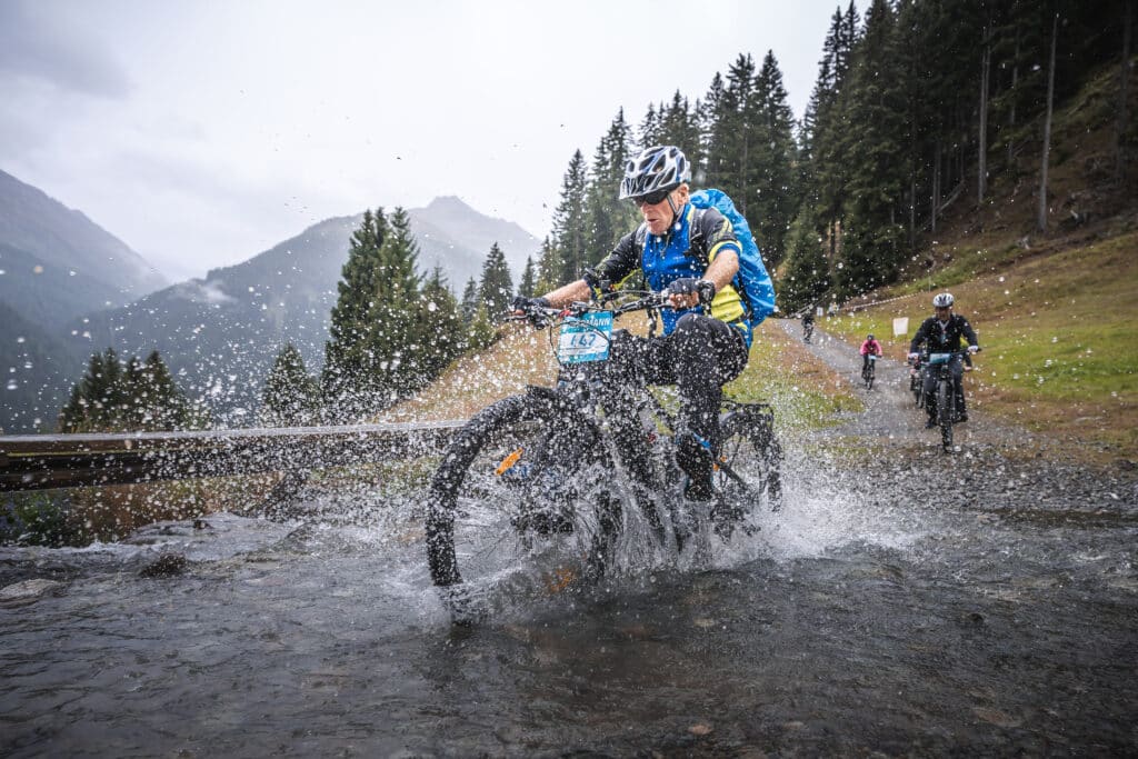 Mann auf e-MTB fährt vor einer Bergkulisse durch eine Pfütze