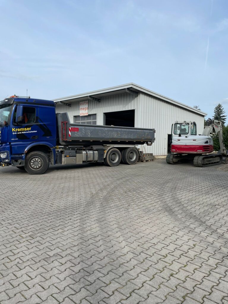 Ein großer LKW mit Ladefläche steht vor einer Halle neben einem Bagger